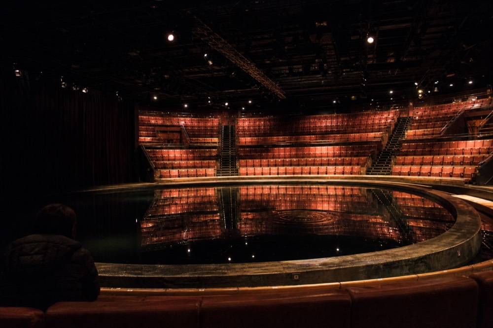 Interior of a large theatre-in-the-round venue with a dark stage pool before a performance