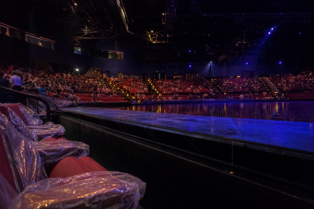 Front rows of a theatre auditorium with waterproof ponchos folded on seat backs beside a stage pool