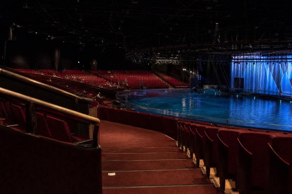 A wide theatre aisle curving down toward a pool stage with empty red seating rows