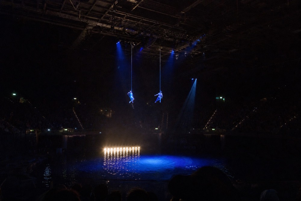 Performers suspended on wires above a dark theatre pool